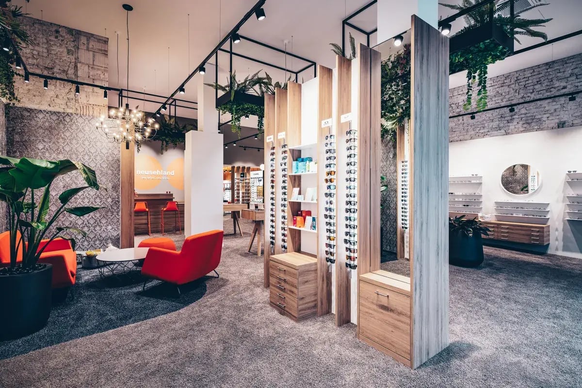 Heller Optikerladen mit Brillenregalen, Hängepflanzen, Pendelleuchten und rotem Sessel.