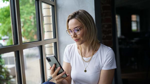 Eine Frau sitzt vor einem Fenster und schaut dabei auf ihr Handy, sie trägt eine Blaulichtfilterbrille